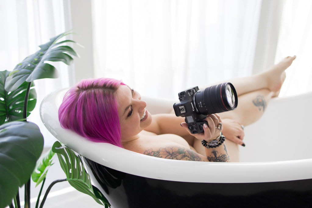 Woman with tattoos lying in a bathtub, smiling with a professional camera in her hand and taking a picture of something off-camera.