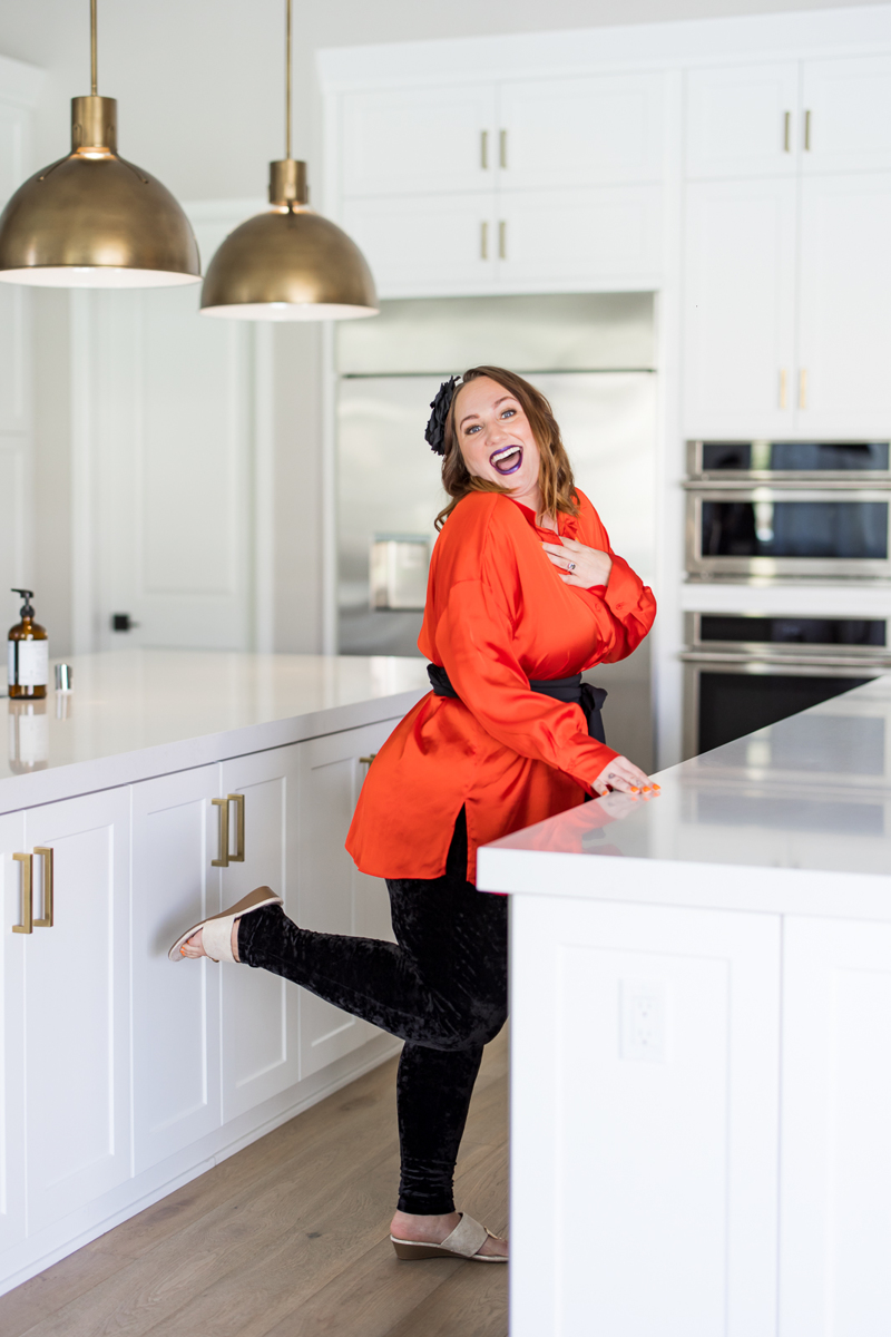 Woman in an orange top and black pants standing in a beautiful white kitchen, smiling while kicking out her leg. 