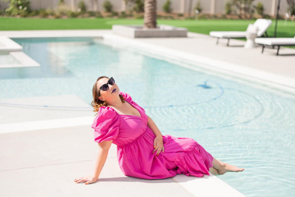 Woman outside by the pool wearing a pink dress with sunglasses as her personal branding outfit, sitting on the ground and posing. 
