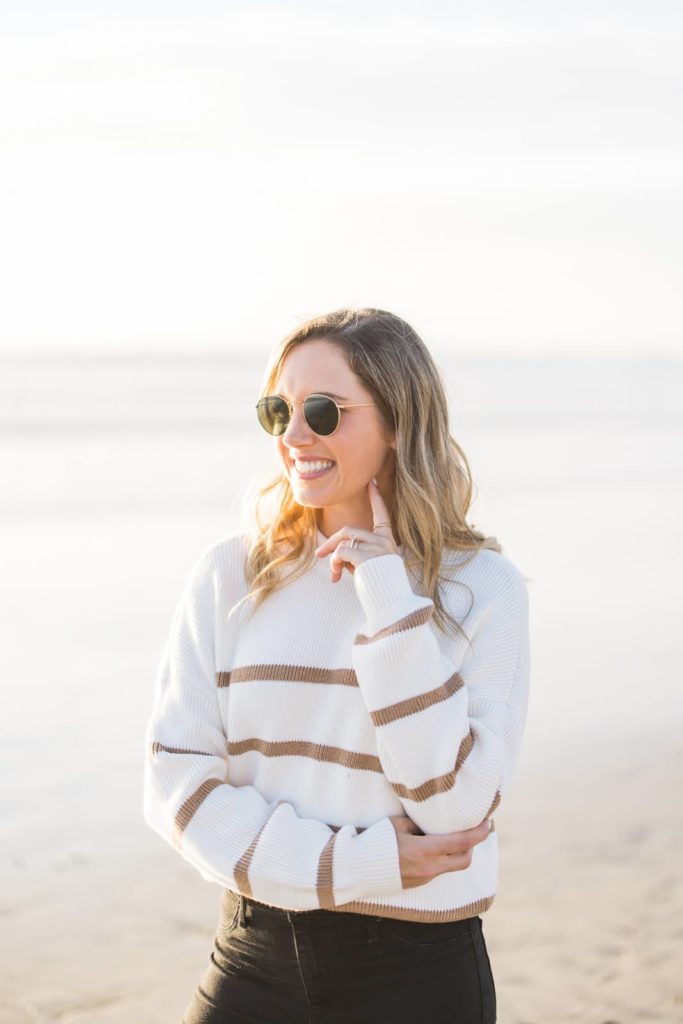 Woman in a white sweatshirt with gold stripes, standing on the San Diego beach and smiling off-camera. 