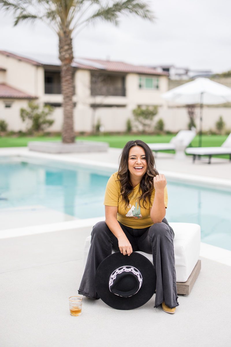 Woman sitting outside by the pool, wearing a gold top with dark gray pants, holding a big hat, and smiling. 