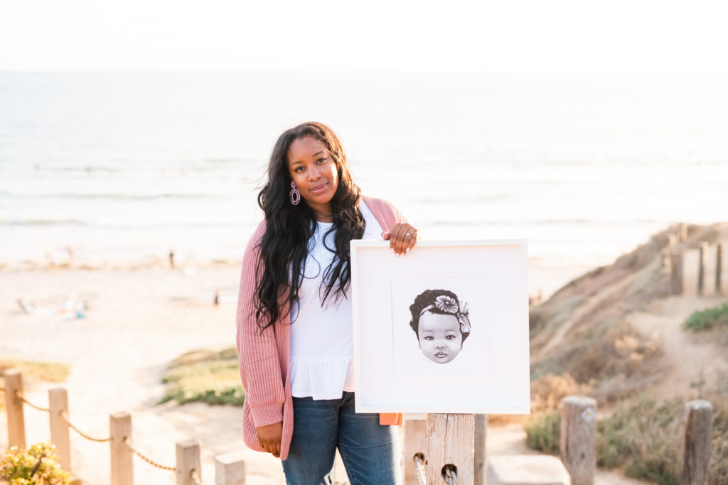 Kia Davis standing at a beach holding a portrait of a little girl who has flowers in her hair. 