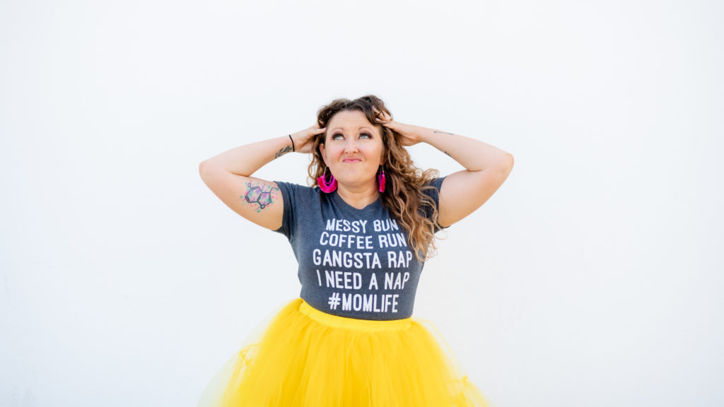  Mompreneur Carly Wright of The Vibrant Mama Method holding her head in excitement and looking up while wearing a mom life t-shirt and bright yellow skirt.  
