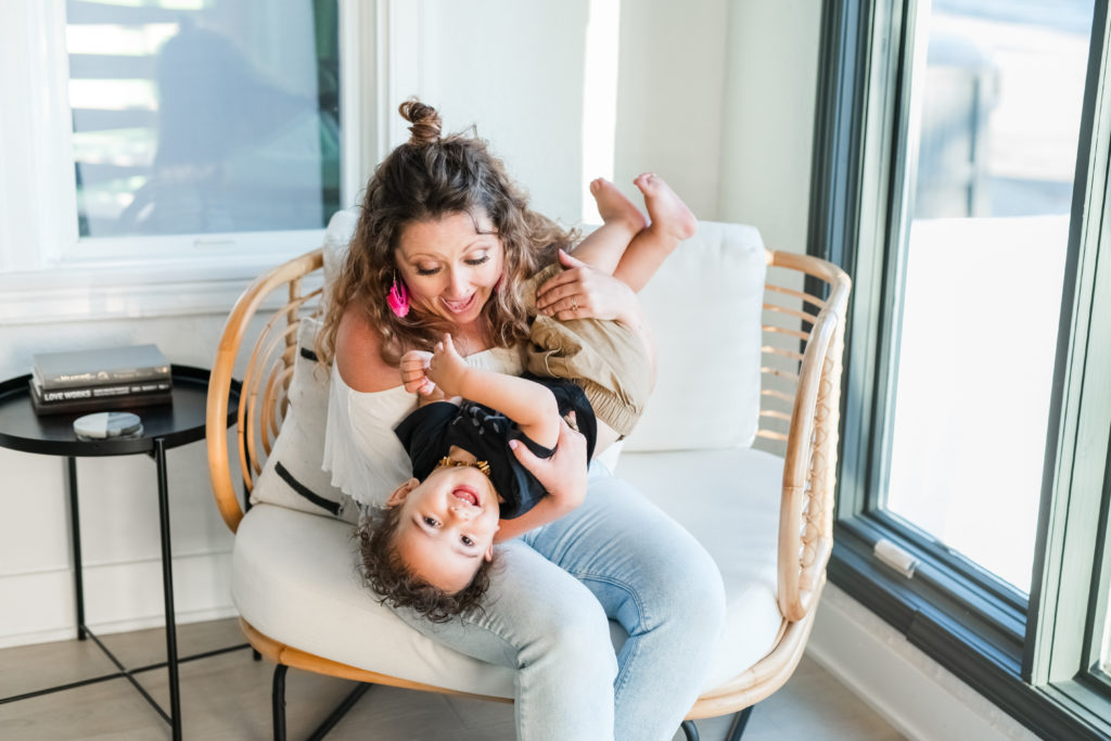 Mompreneur Carly Wright sitting in a chair while holding her son upside down, playing and laughing with him. 