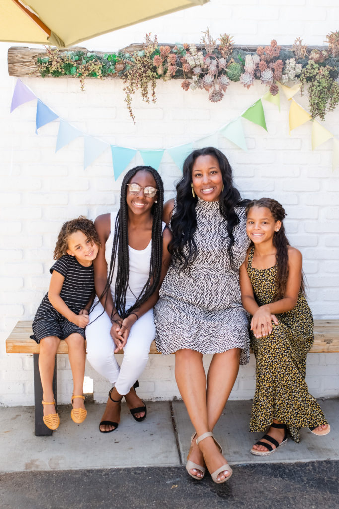 Kia Davis standing in front of a pink wall with her three daughters while they all smile and have fun. 

