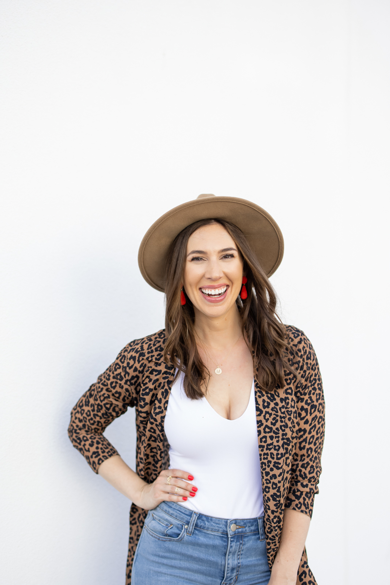  Woman in leopard print and a hat, with one hand on her hip, smiling at the camera during her branding photography session.   
