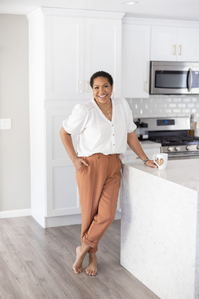 Woman standing in kitchen in neutral colors with flowing pants and white top with big smile. 