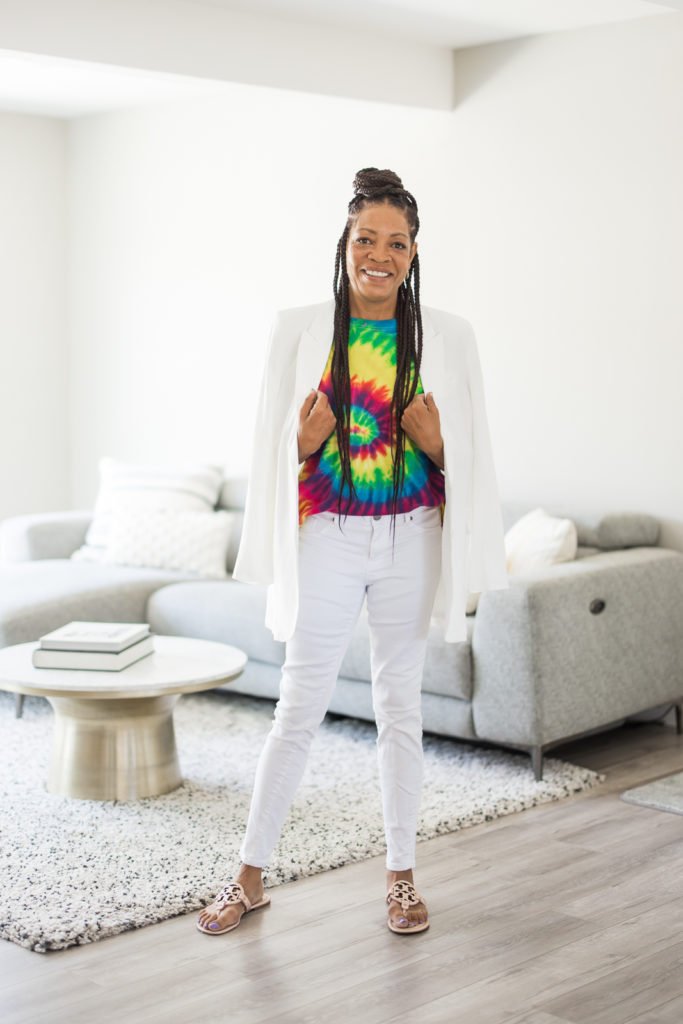Woman in a tie-dye shirt and blazer, standing in the living room, looking at the camera.