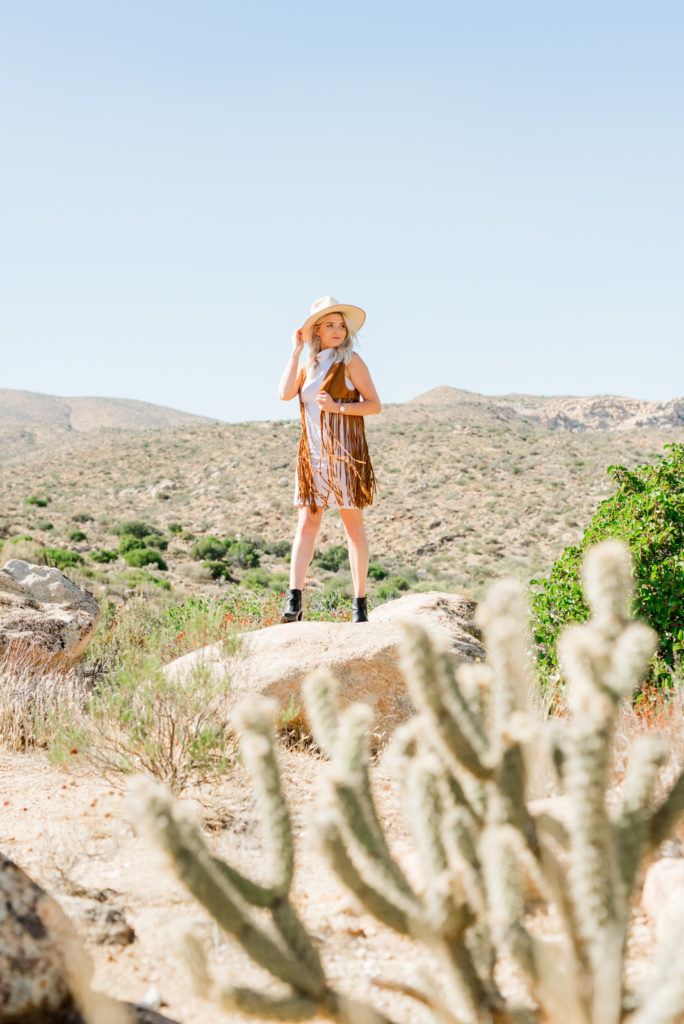 Woman standing in the desert on top of a rock wearing a big hat, and looking away from the personal brand photographer. 