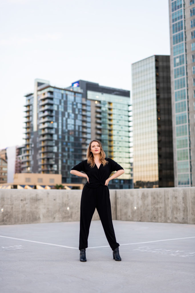 Woman standing on a rooftop wearing all black with big buildings behind her. 