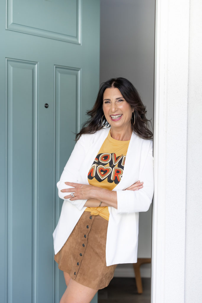 Woman standing in a doorway wearing a white jacket and a brown skirt while smiling. 