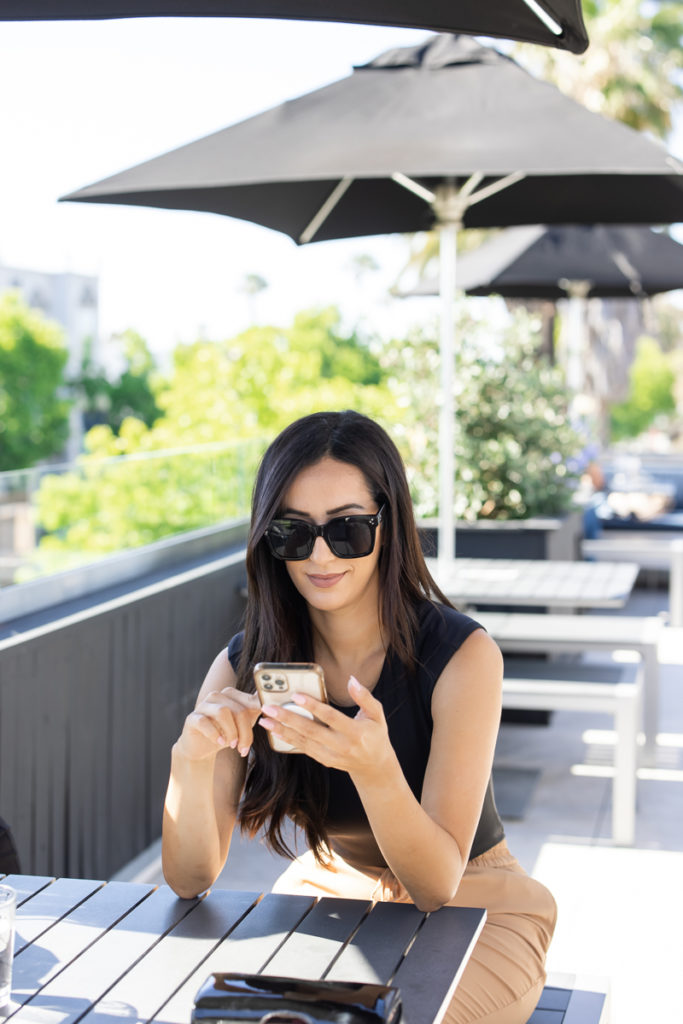 Jenica Martin sitting at an outside cafe with sunglasses on, looking at her phone. 

