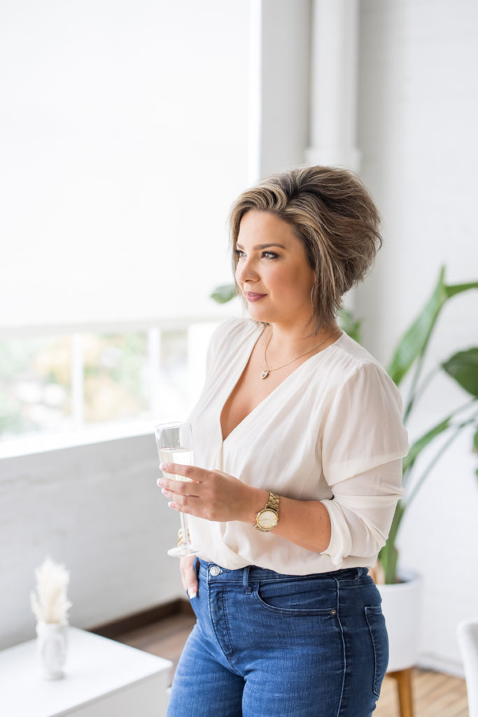 Woman in a white top and jeans holding a glass of wine and looking away from the camera.