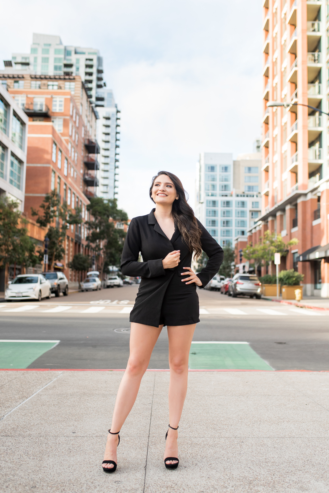 Woman standing in the city of San Diego, surrounded by buildings, wearing all black with black heels. 

