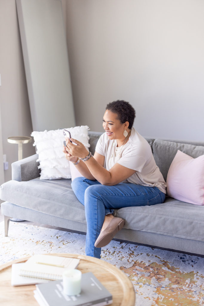 Woman sitting on the couch with her phone in front of a coffee table smiling during her brand shoot. 
