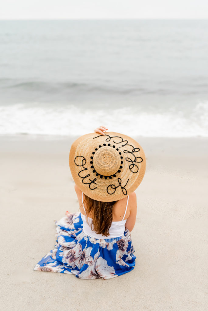 The back of a woman sitting on the beach with a big hat that says out of office on the top.
