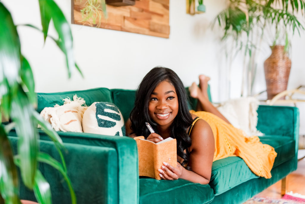 Woman journaling about her money mindset goals while lying on a green couch and smiling. 