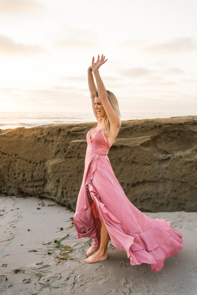 Woman in a pink dress at the San Diego beach with her hands in the air and smiling. 
