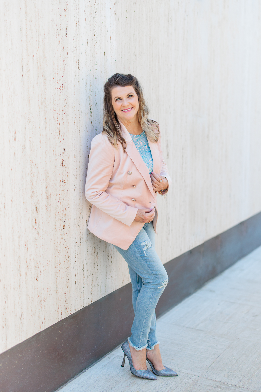 Woman with jeans and a pink blazer standing on an outside wall smiling at the camera. 
