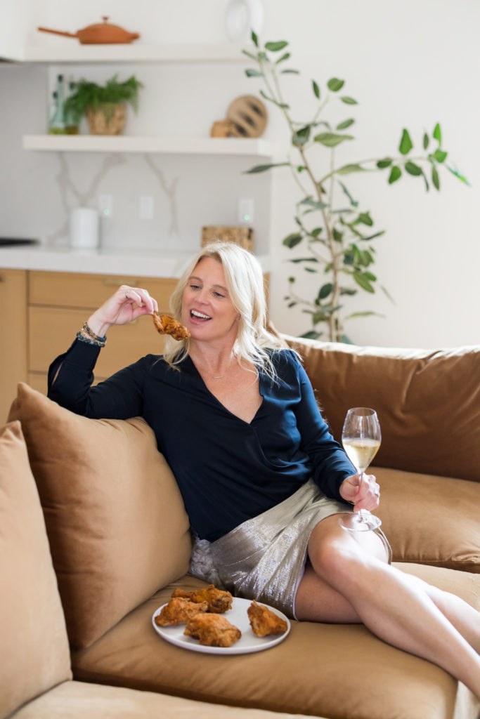 Woman sitting on the couch eating chicken and drinking wine.