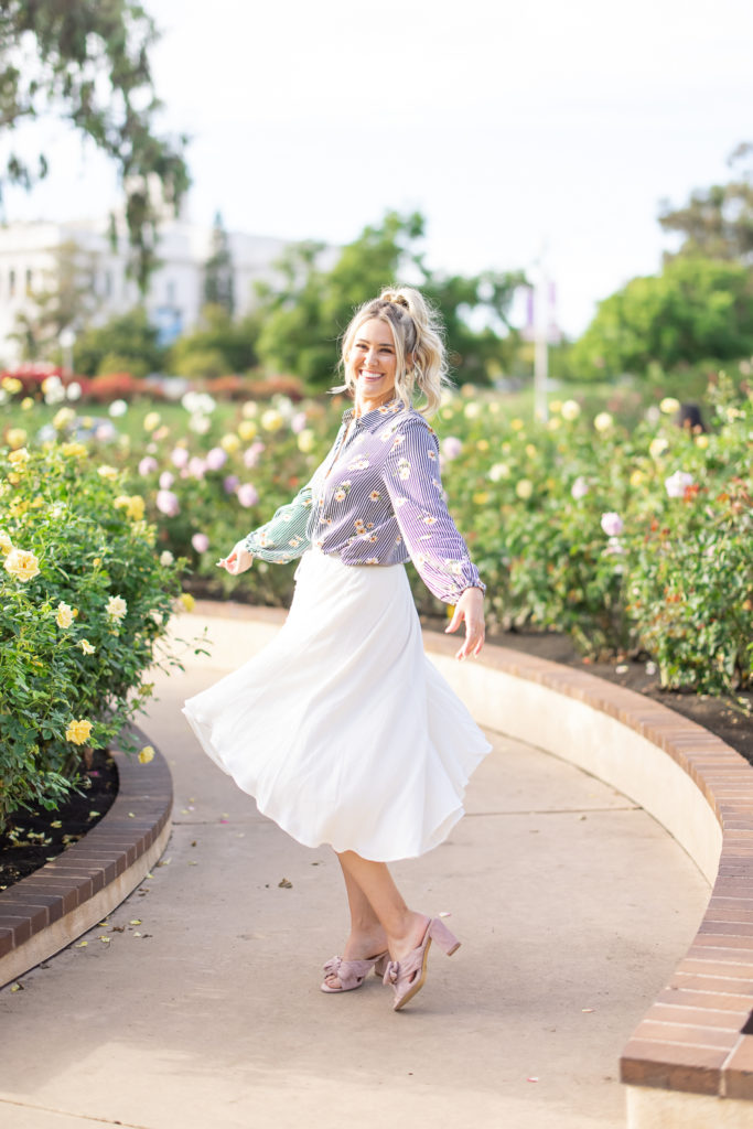Woman twirling around in a garden wearing a white skirt and floral blouse. 