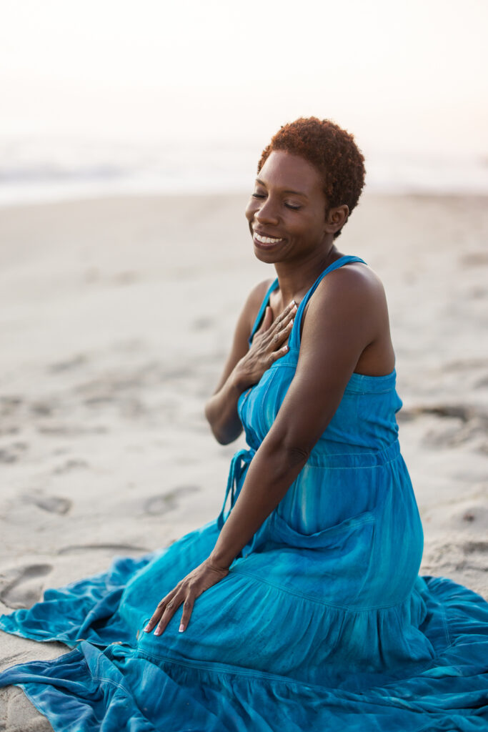 Woman in a blue dress on the San Diego sand with her hand to her heart and smiling.
