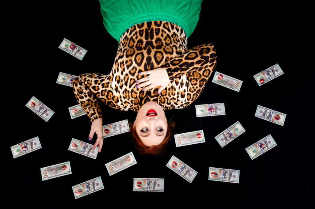 Woman in a leopard top and green bottoms lying on the floor surrounded by money.