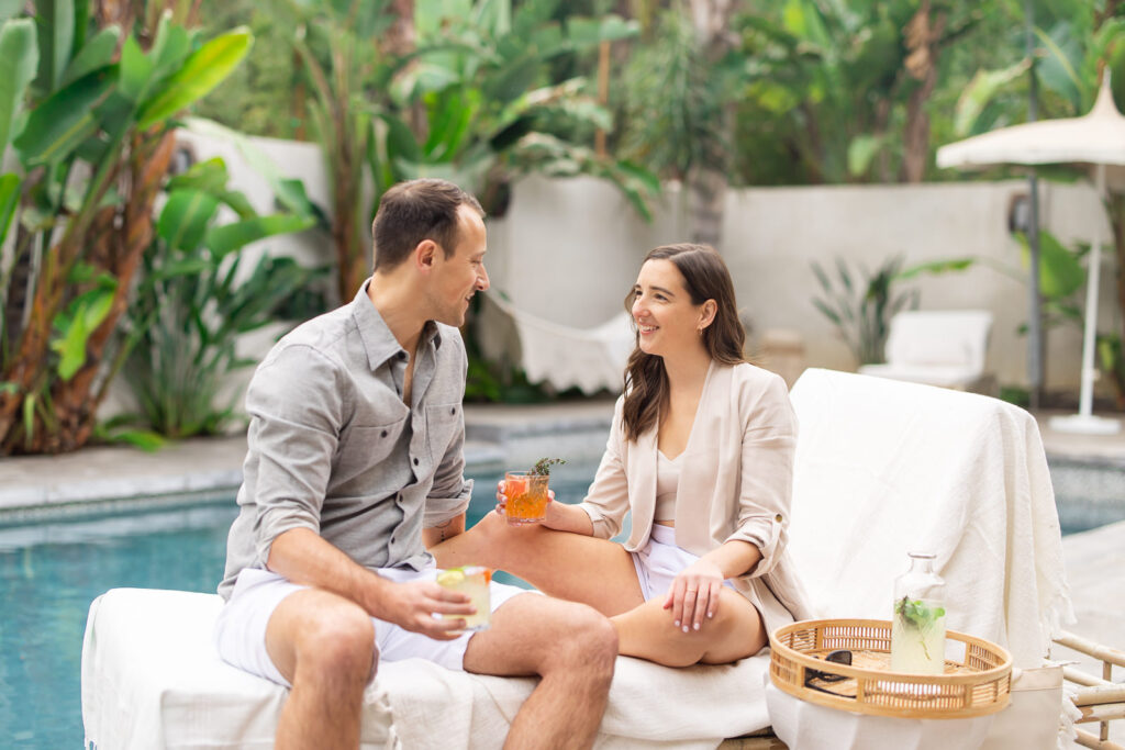 Krystle holding a drink and sitting on a pool lounge chair next to her husband and smiling at him.