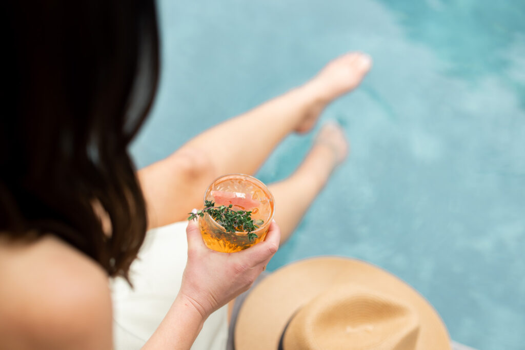 The back of a woman sitting next to the pool with her feet in the water as she holds a drink.