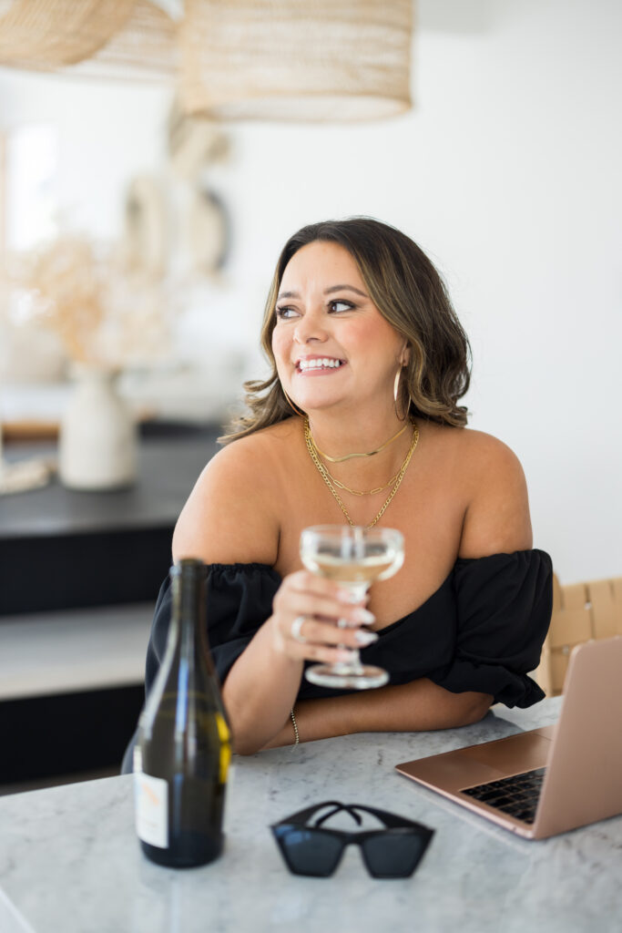 Woman wearing a black off the shoulder shirt, three thing gold necklaces while holding a glass with champagne and looking away