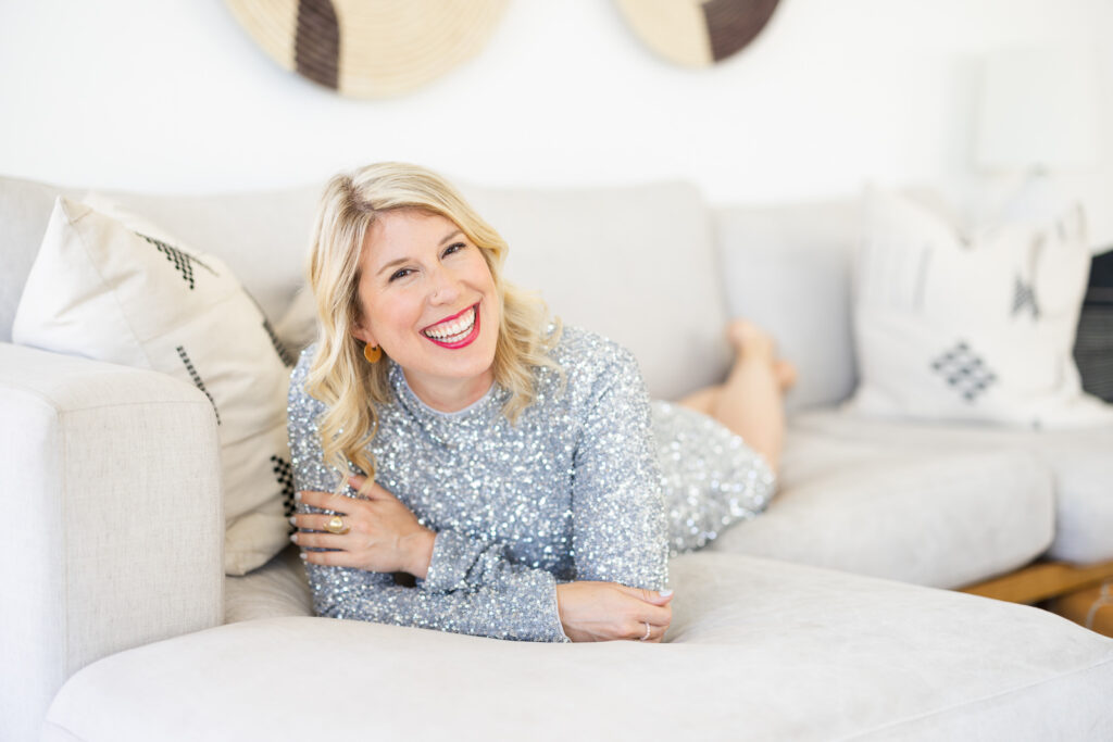 Woman wearing a glittery, silver outfit lying on a white couch and smiling during her mini session for photography. 
