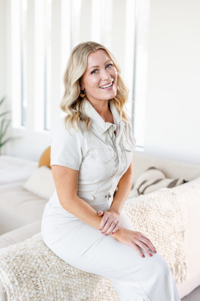 Meg wearing a white jumpsuit and sitting on the top part of a couch smiling with her legs crossed. 