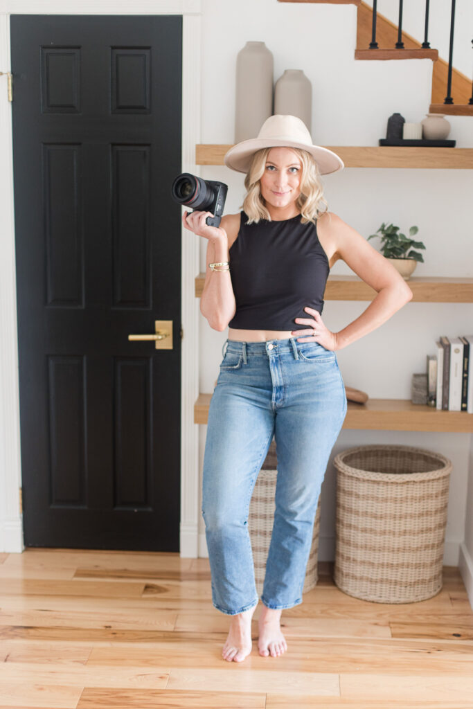 Meg Marie wearing a crop top, jeans, and a hat, posing with one hand holding a camera and the other hand on her hip.

