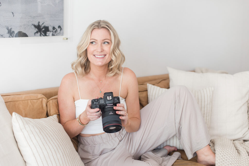 Meg Marie wearing tan pants and a white crop top, sitting on a couch smiling and holding her camera.