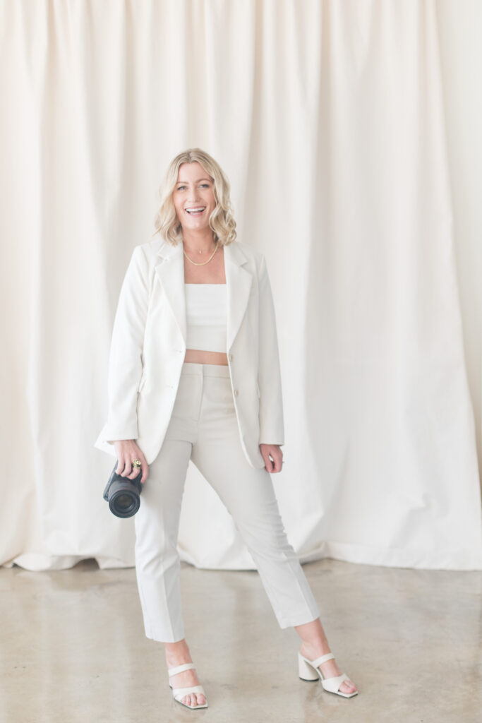 Photography business coach Meg Marie wearing all white holding a camera in one hand and smiling. 
