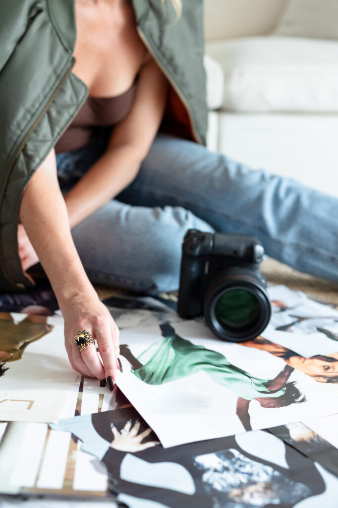 Woman in jeans, green jacket, and brown top sitting on the floor with a camera and printed photo collection, capturing her creative brand process.