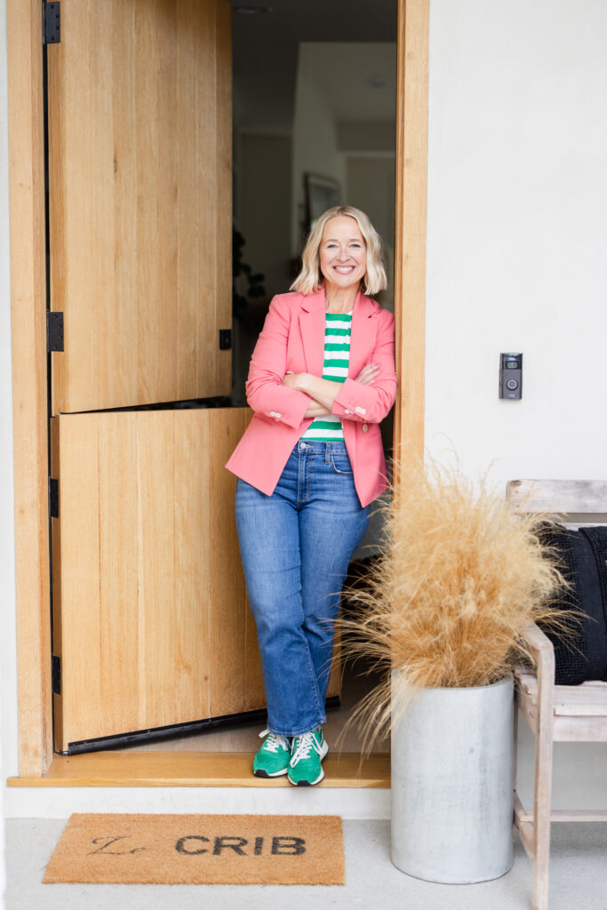 Woman leaning on a door frame wearing a pink blazer, white and green striped shirt, and green sneakers—blending casual style with confident brand presence.