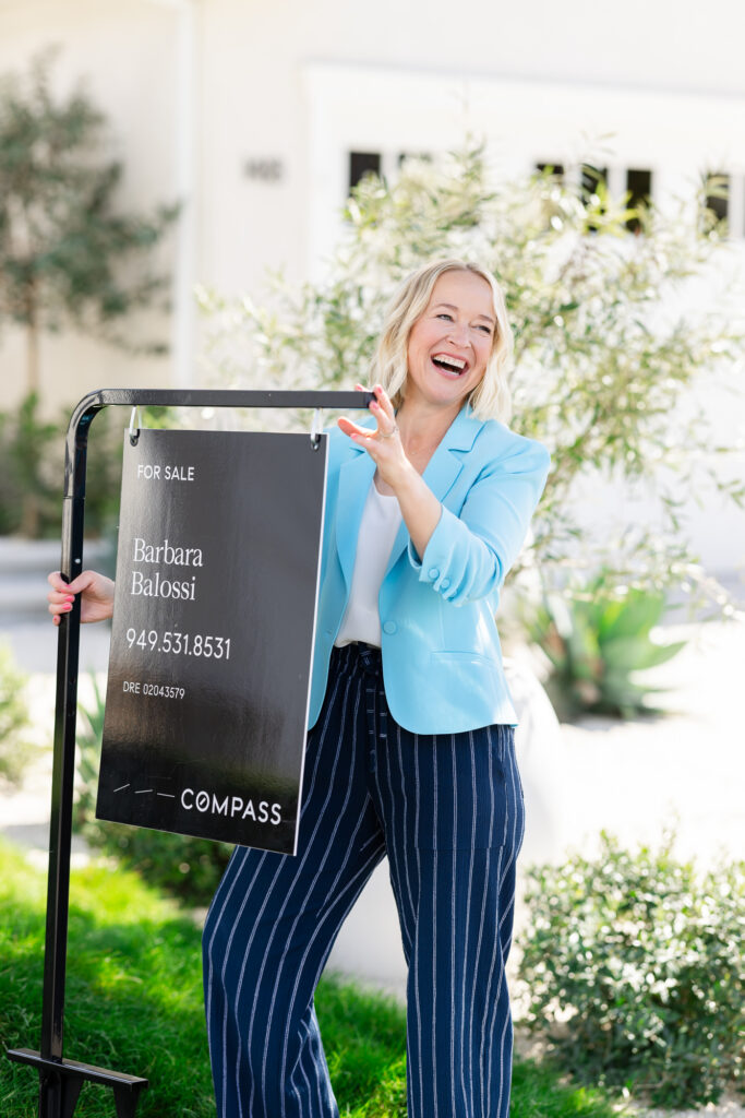 Female real estate educator in a light blue blazer, white blouse, and navy striped pants holding a sign—capturing a confident, trustworthy image for personal real estate branding.