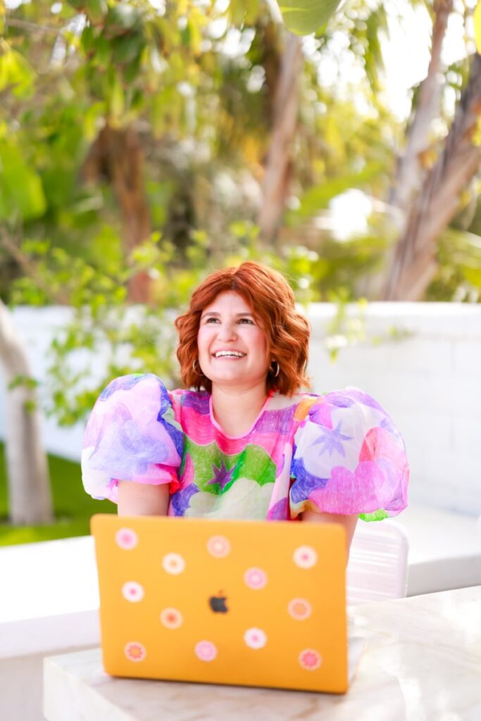 A person with red hair smiles while seated behind a yellow laptop covered with stickers. They wear a colorful, puffy-sleeved top. Green foliage is visible in the background.
