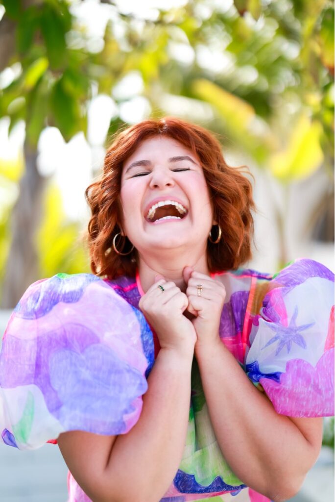 A woman with red hair joyfully laughs with eyes closed, hands clasped under her chin. She's wearing a colorful, vibrant dress with puffed sleeves.