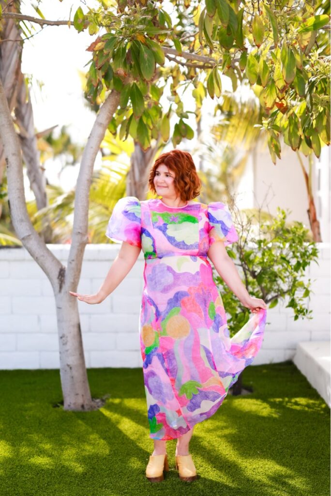 A woman with red hair wears a colorful, patterned dress and yellow shoes, standing on lush green grass. She is under a tree and looks content.
