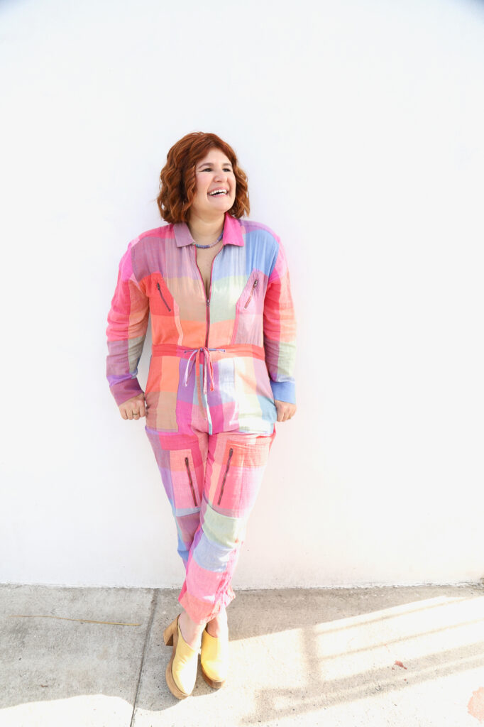 Smiling woman in a colorful patchwork jumpsuit leans against a white wall. She exudes joy and confidence, wearing yellow clogs on a sunny day.