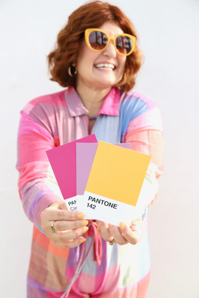 Woman in colorful attire holding three Pantone color cards in pink, purple, and yellow. She smiles joyfully, exuding a playful, vibrant mood.