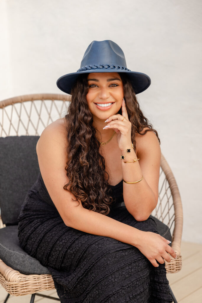 A woman with long curly hair, wearing a blue hat and black dress, smiles warmly while seated on a wicker chair. She exudes a relaxed, confident aura.