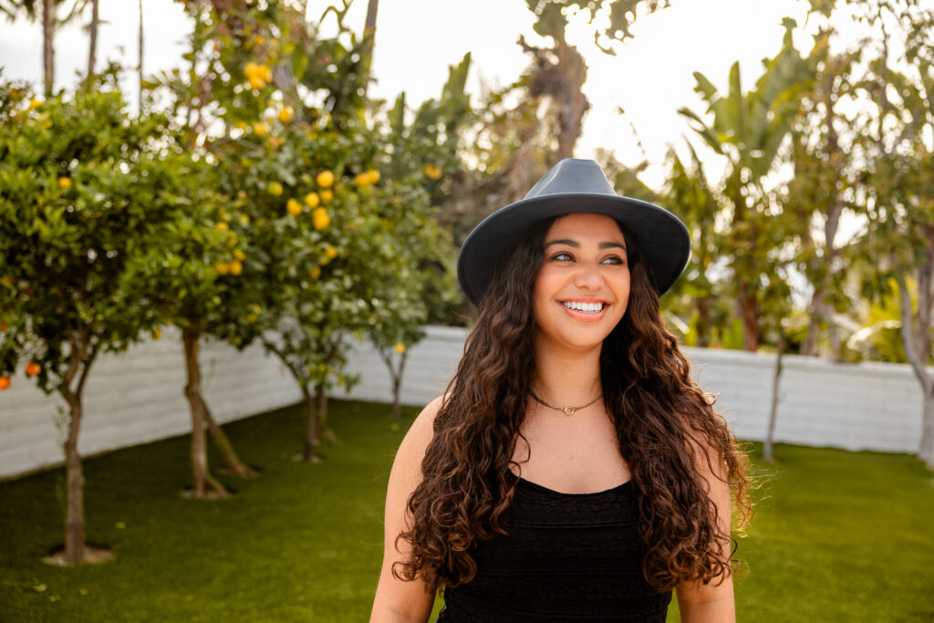 Smiling woman with long curly hair and hat stands in a sunny garden with lush green grass. Lemon trees and a white fence create a serene backdrop.