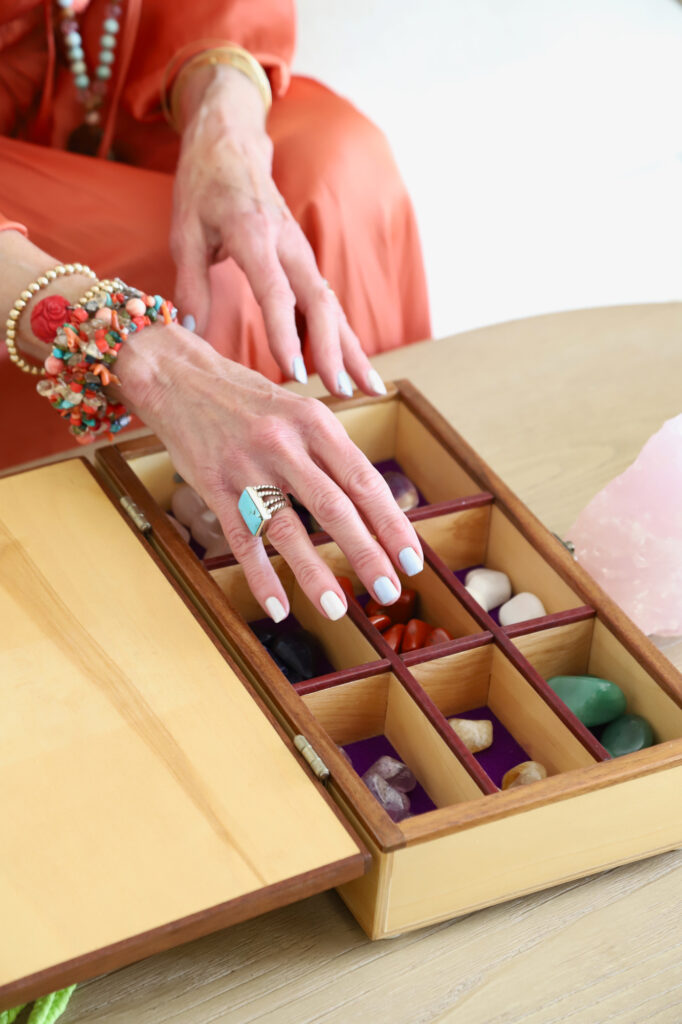 A hand, adorned with colorful bracelets and a turquoise ring, reaches into a wooden box filled with various crystals, conveying a sense of calm and spirituality.