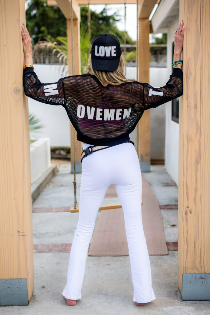 Woman standing with back to camera, wearing a black "LOVE" cap, mesh top with "MOVEMENT," and white pants. She leans on wooden beams, conveying confidence.