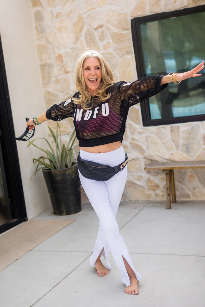 A woman joyfully poses with arms outstretched, wearing a mesh top with "MINDFUL" text, white pants, and a waist bag. Background shows a stone wall, plant, and window.