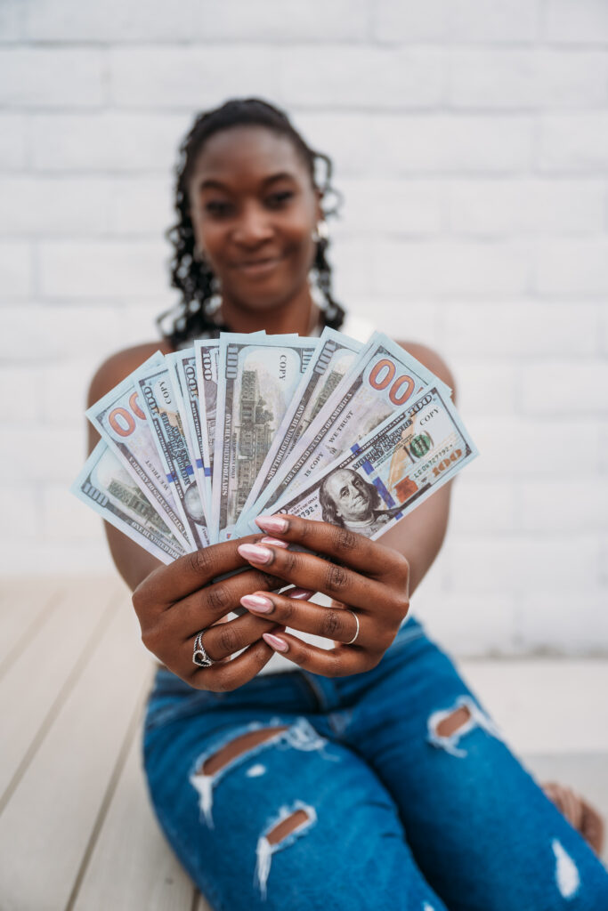 A person in ripped jeans holds out a fan of hundred-dollar bills with a confident smile. The background is a blurred white brick wall.