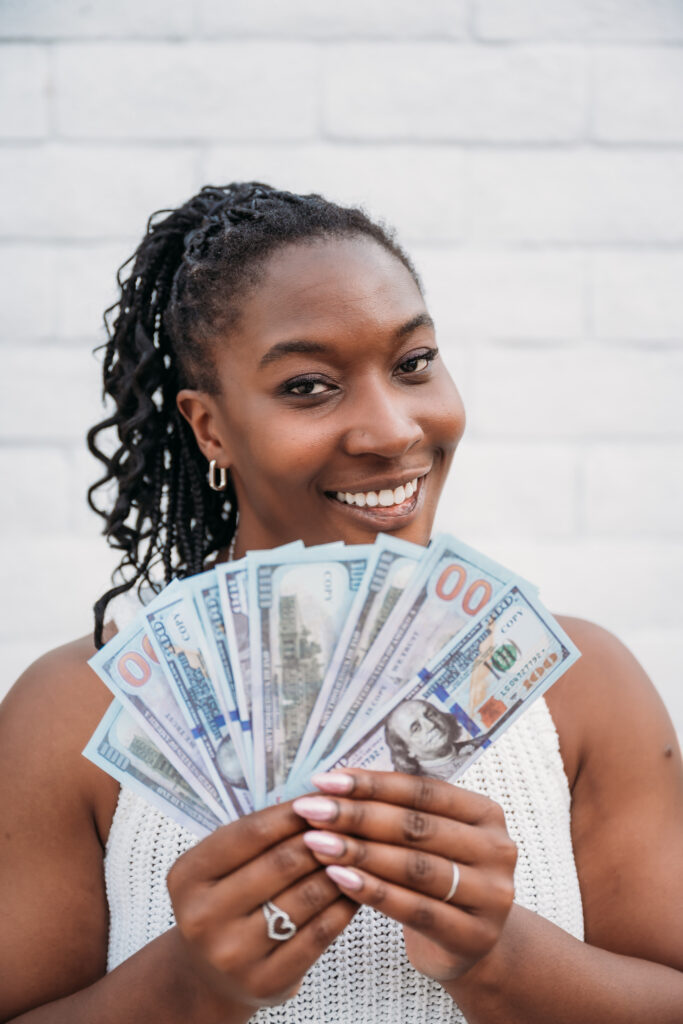 Smiling woman in a white top holds a fan of hundred-dollar bills. She's in front of a white brick wall, conveying a sense of happiness and success.
